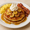 Stack of pancakes with bananas, syrup, whipped cream, scrambled eggs, and bacon on a white plate. halo bagels harlem