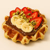Waffle with Nutella, sliced bananas, strawberries, and pistachios on a beige background harlem halo bagels